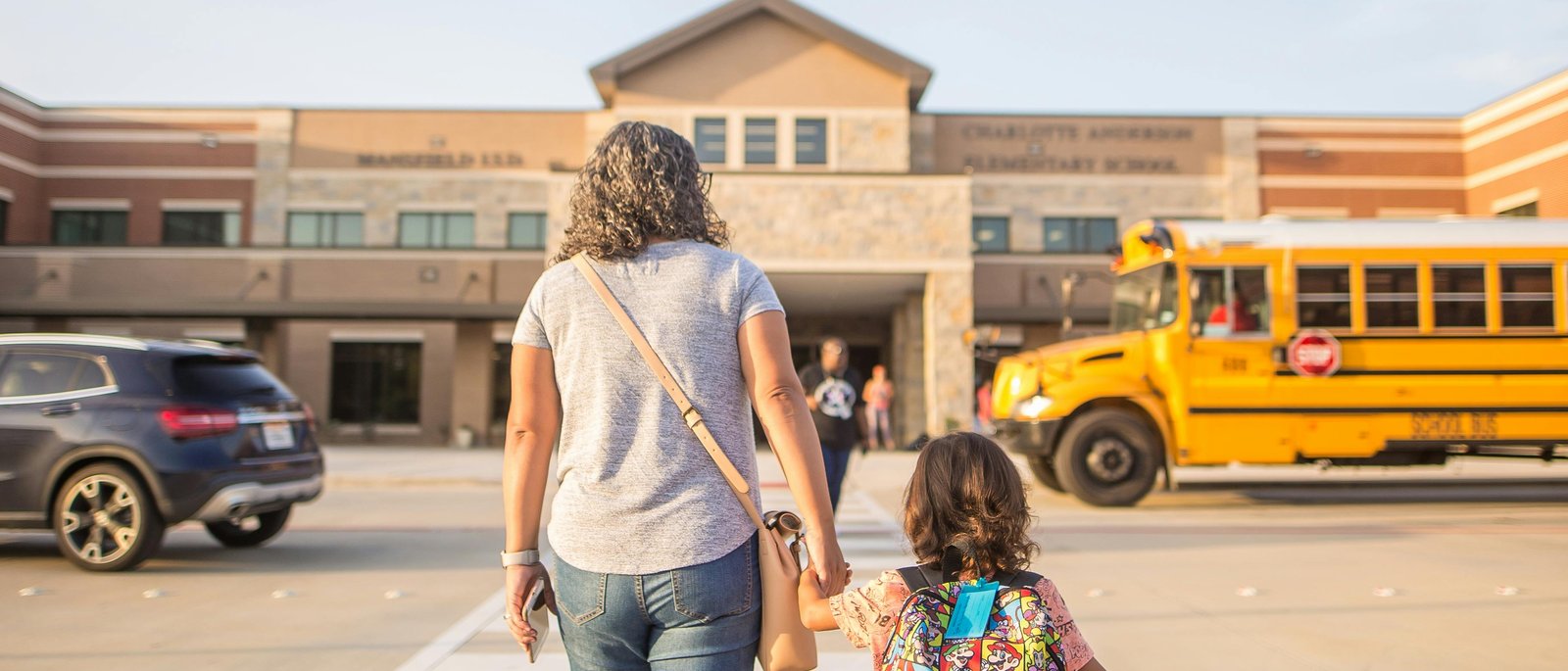 Parent walking child to elementary school, with school bus in view